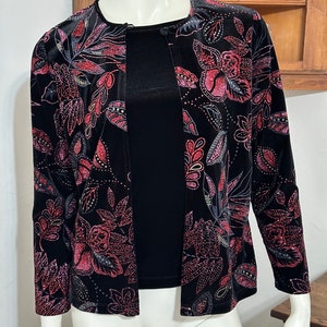 May include: A black velvet jacket with a floral pattern in shades of pink and gray. The jacket has a button closure and long sleeves.