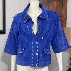 May include: A vibrant royal blue suede jacket featuring three-quarter length sleeves, a fold-over collar, and a front zipper closure. The jacket is accented with silver-toned metal studs along the collar, front placket, and two front pockets.