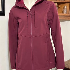 May include: A burgundy softshell jacket with a zipper closure and a hood. The jacket has a zippered pocket on the left chest and two zippered pockets on the lower front. The jacket is made by the brand "Lands' End".