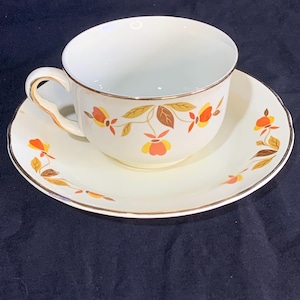 May include: A white teacup and saucer with a gold rim and a floral pattern in shades of orange, yellow, and brown. The pattern features stylized leaves and flowers.