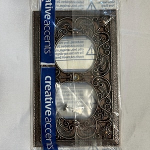 May include: A decorative bronze-colored double electrical outlet cover with an ornate floral design. The cover is packaged in clear plastic and has the words "creative accents" printed on a blue strip.