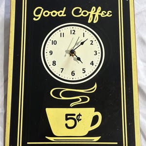 May include: A rectangular black and yellow wall clock featuring the text Good Coffee at the top. The clock face has black numbers and hands. Below the clock is a yellow coffee cup graphic with the text 5 cents and steam rising above it.