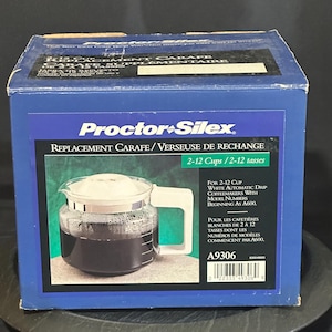 May include: A blue box with a Proctor-Silex replacement carafe. The box features a picture of the glass carafe with a white handle and lid, filled with dark liquid. Text on the box reads "REPLACEMENT CARAFE/VERSEUSE DE RECHANGE".