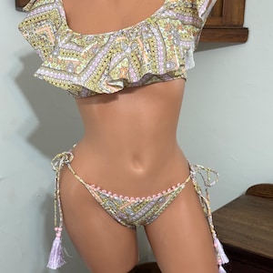 May include: A white and yellow patterned bikini top with a ruffled neckline and a matching bikini bottom with pink trim and lavender tassels.