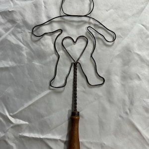 May include: A dark metal wire frame shaped like a teddy bear with a heart-shaped cutout in the center. The wire frame is attached to a twisted metal rod that connects to a smooth, cylindrical wooden handle at the base of the object.