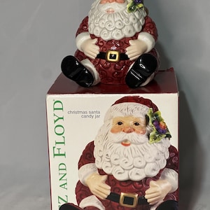 May include: A ceramic Christmas Santa candy jar with a red suit, black boots, and a white beard. The jar has a gold buckle and is sitting on a box with the text "Fitz and Floyd". The Santa figure has a festive hat with a floral accent.