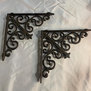 May include: Two dark brown metal shelf brackets featuring an intricate scrollwork design. Each bracket has a triangular shape with a flat top and side edge for mounting, and a decorative openwork pattern of swirling curves and pointed accents.