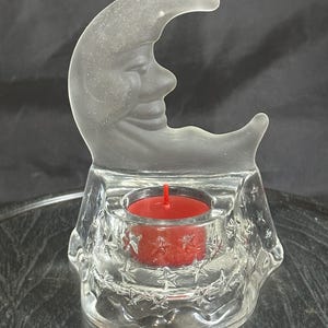 May include: A clear glass tea light candle holder featuring a frosted crescent moon with a face at the top and a base decorated with embossed stars. A small red tea light candle is placed in the center circular opening of the glass holder.
