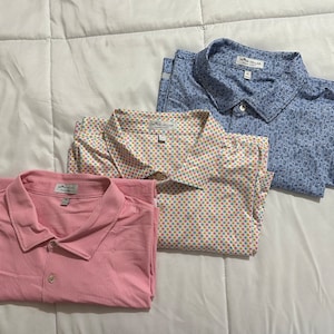 May include: Three men's short-sleeved shirts with button-down collars. The shirts are in different patterns: pink with a subtle texture, white with colorful polka dots, and blue with a small, abstract pattern. The shirts all have the same brand label: "Peter Millar, Classic Fit".
