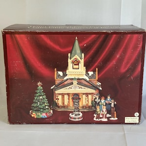 May include: A Christmas village scene with a church, Christmas tree, and figurines. The church has a green steeple and is decorated with snow. The Christmas tree is green with colorful presents. The figurines depict people in winter attire.