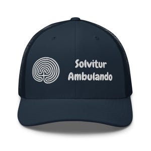 May include: A navy blue trucker cap featuring a white embroidered labyrinth symbol and the Latin phrase "Solvitur Ambulando". The cap has a mesh back and a curved peak, suitable for various outdoor activities.