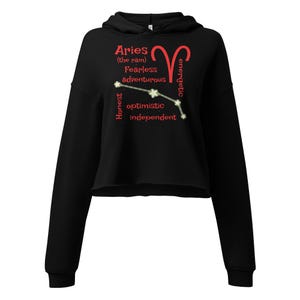 May include: Black cropped hoodie with the word "Aries" and a red ram symbol. The hoodie features the words "Fearless, adventurous, energetic, honest, optimistic, independent" in red. The hoodie has long sleeves and a hood.