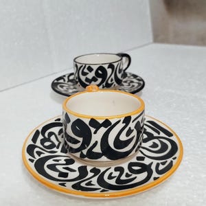 Puede incluir: Dos tazas de café de cerámica blanca con diseños de caligrafía árabe negra y ribete naranja. Las tazas están sobre platillos a juego.