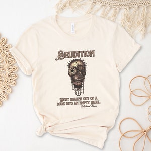 May include: A beige t-shirt with a brown and black graphic of a skull with gears and the text "ERUDITION" above it. The text below the graphic reads "Just shaken out of a book into an empty shell. - Voltaire"