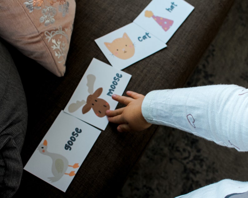 May include: A child's hand points to a flashcard with a brown moose illustration and the word "moose" written vertically in blue letters. Other flashcards with illustrations of a goose, cat, and hat are visible on a brown surface.