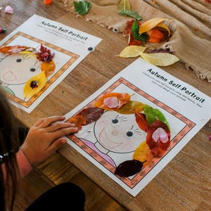 May include: Two "Autumn Self-Portrait" art projects with leaf hair are displayed on a wooden table. The artwork features a drawn face with a leaf crown. Crayons and colorful leaves are scattered around the projects.