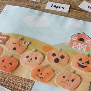 Puede incluir: Un conjunto de nueve pegatinas de fieltro naranja en forma de calabaza con diferentes expresiones faciales, incluyendo feliz, triste, dormido y sorprendido. Las pegatinas est&aacute;n dispuestas sobre un fondo marr&oacute;n con un cielo azul y un granero rojo en el fondo. El texto "Pumpkin Patch" es visible en un cartel marr&oacute;n.