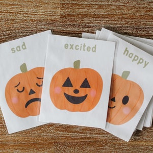 Puede incluir: Tres tarjetas blancas con ilustraciones de calabazas naranjas y las palabras "triste", "emocionado" y "feliz" impresas en ellas.