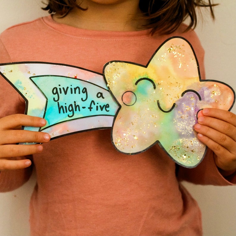 Puede incluir: Un ni&ntilde;o sostiene una estrella de papel colorida con una cara sonriente y una cola de arco&iacute;ris que dice "giving a high-five".