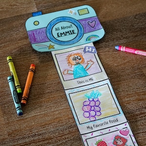 Op de afbeelding: Een papieren knutselcamera met de tekst All About EMMIE, voorzien van een uittrekbare strook met handgetekende illustraties waaronder een persoon met het label This is ME, een paarse druiventros met het label My favorite food en een aardbei, vergezeld van drie kleurpotloden.