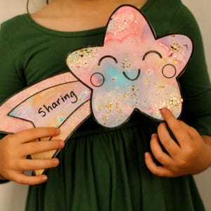 Puede incluir: Un ni&ntilde;o sostiene una estrella rosa y azul con brillantina y una cara sonriente. La estrella est&aacute; unida a un arco&iacute;ris rosa y blanco con la palabra "compartir" escrita en ella.