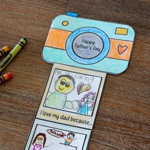 May include: A Father's Day card in the shape of a camera, with the words "Happy Father's Day." The card includes drawings and the phrases "He is caring" and "I love my dad because..." Crayola crayons are scattered around the card.