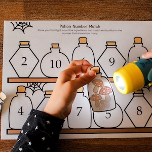 May include: A Potion Number Match educational activity sheet featuring outlines of potion bottles with numbers and a shelf design. A small flashlight illuminates a cut-out potion bottle card containing illustrations of five red and white mushrooms.