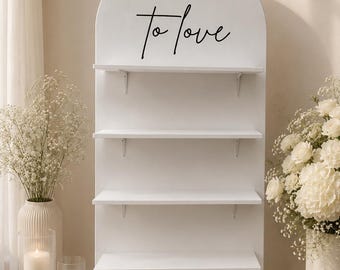 May include: A white arched display shelf featuring four horizontal shelves and a base shelf. The top of the arch is printed with the black text CHEERS! to love. The structure is made of smooth white material with visible support brackets.