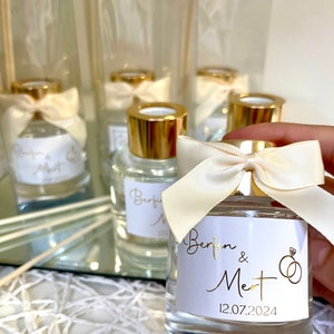 Könnte beinhalten: Ein Set kleiner Glasflaschen mit goldenen Deckeln und weißen Etiketten. Die Etiketten tragen die Namen "Berfin & Mert" und das Datum "12.07.2024" in goldener Schrift. Jede Flasche ist mit einem weißen Satinband um den Hals gebunden.
