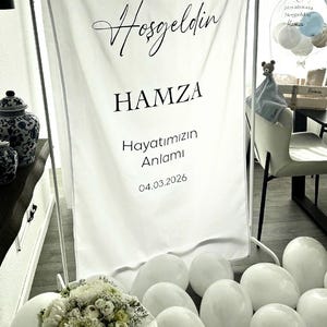 May include: A white fabric banner on a metal stand featuring the black text Hosgeldin HAMZA Hayatımızın Anlamı 04.03.2026. The banner is surrounded by white balloons and a bouquet of white flowers, creating a decorative display for a celebration.