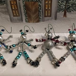May include: Four handmade Christmas ornaments in the shape of whimsical figures. Each ornament features a silver head, beaded body, and wire arms. The ornaments have accents of blue, silver, and red, with a wire loop for hanging. The background is a snowy winter scene.