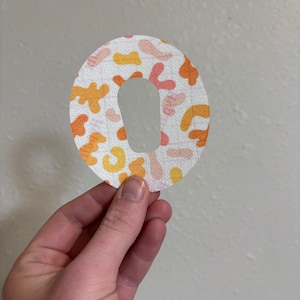 May include: A white, round, adhesive bandage with a hole in the center. The bandage has a colorful abstract pattern in shades of orange, yellow, and pink.