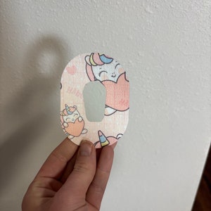 May include: A patterned medical patch featuring a light pink background with cartoon unicorns and hearts. The patch has a curved top and a cutout for a medical device. The unicorn design includes pastel colors.