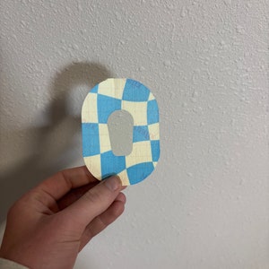 May include: A blue and white checkered oval-shaped patch with a hole in the center. The patch is made of fabric and has a slightly textured surface.
