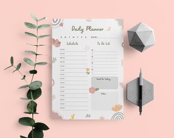 Puede incluir: Un planificador diario con un diseño floral. El planificador tiene secciones para la agenda, la lista de tareas, los objetivos del día y las notas. El planificador está impreso en papel blanco con un fondo gris claro.