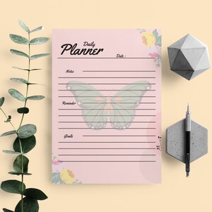 May include: A pink daily planner with a floral design and a butterfly illustration. The planner has sections for notes, reminders, and goals.