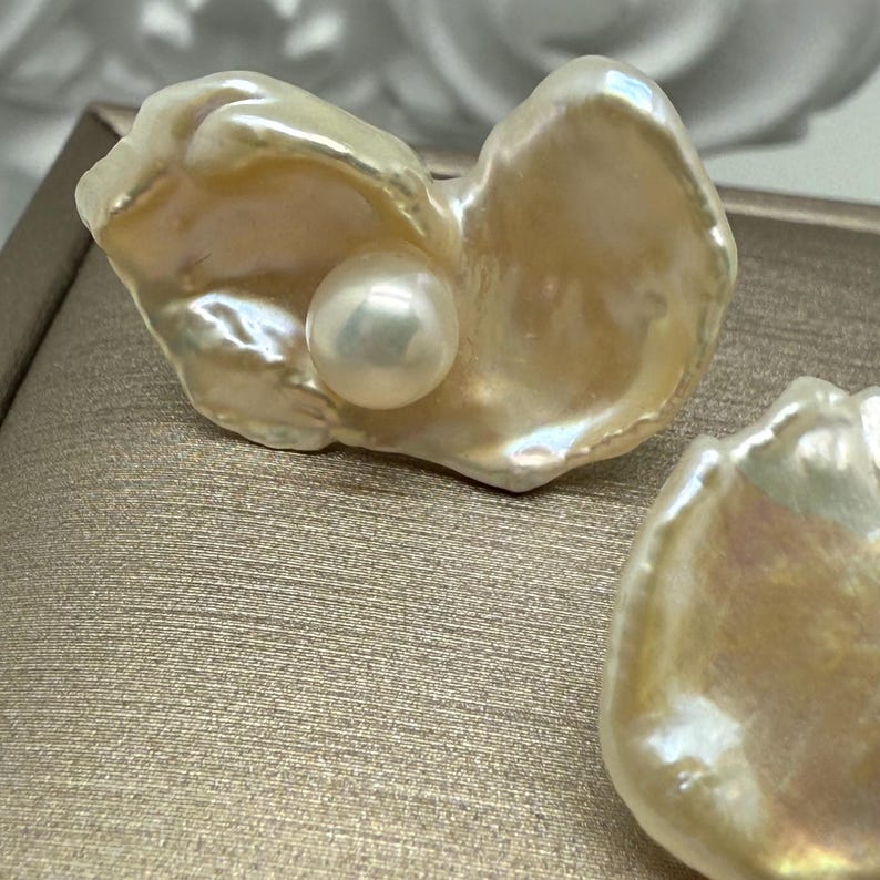 May include: Close-up of a pair of pearl earrings. Each earring features a lustrous, off-white pearl nestled within a creamy, irregular-shaped shell. The shells have a textured surface and a subtle, iridescent sheen, reflecting light. The earrings are set against a neutral background.