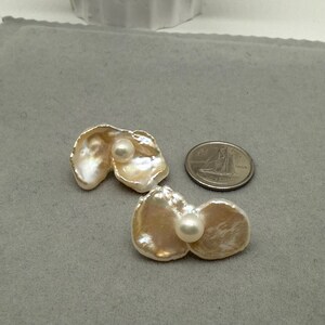 May include: Two pearl earrings with a unique, organic shape. Each earring features a large, iridescent, petal-like form with a smaller, round pearl nestled in the center. The earrings are displayed on a gray surface next to a coin.