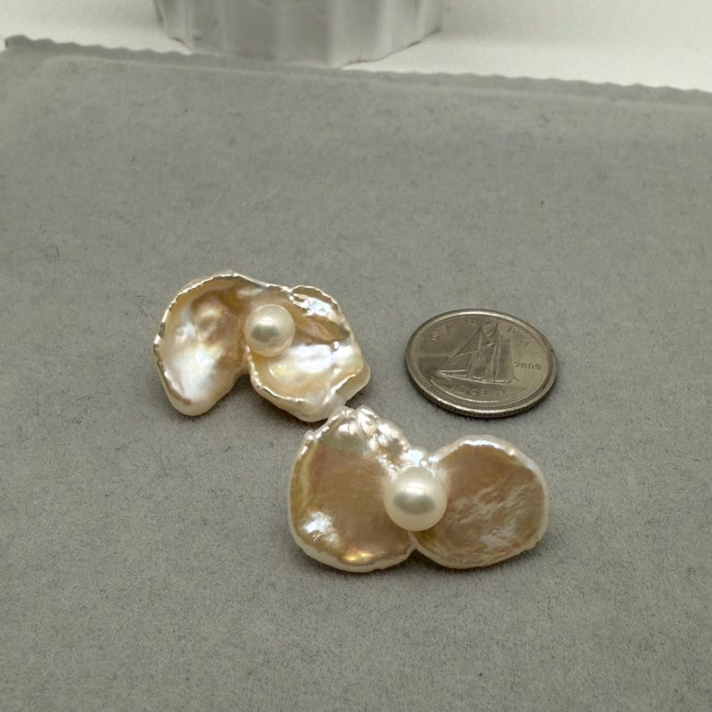 May include: Two pearl earrings with a unique, organic shape. Each earring features a large, iridescent, petal-like form with a smaller, round pearl nestled in the center. The earrings are displayed on a gray surface next to a coin.