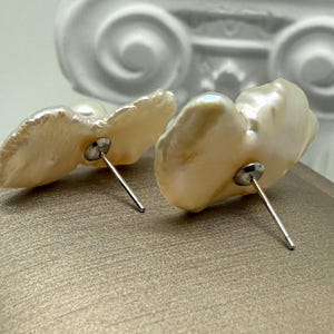 May include: A pair of pearl stud earrings. The earrings feature irregular, off-white pearls with a lustrous sheen. Each pearl is mounted on a silver post. The background is a neutral, textured surface.