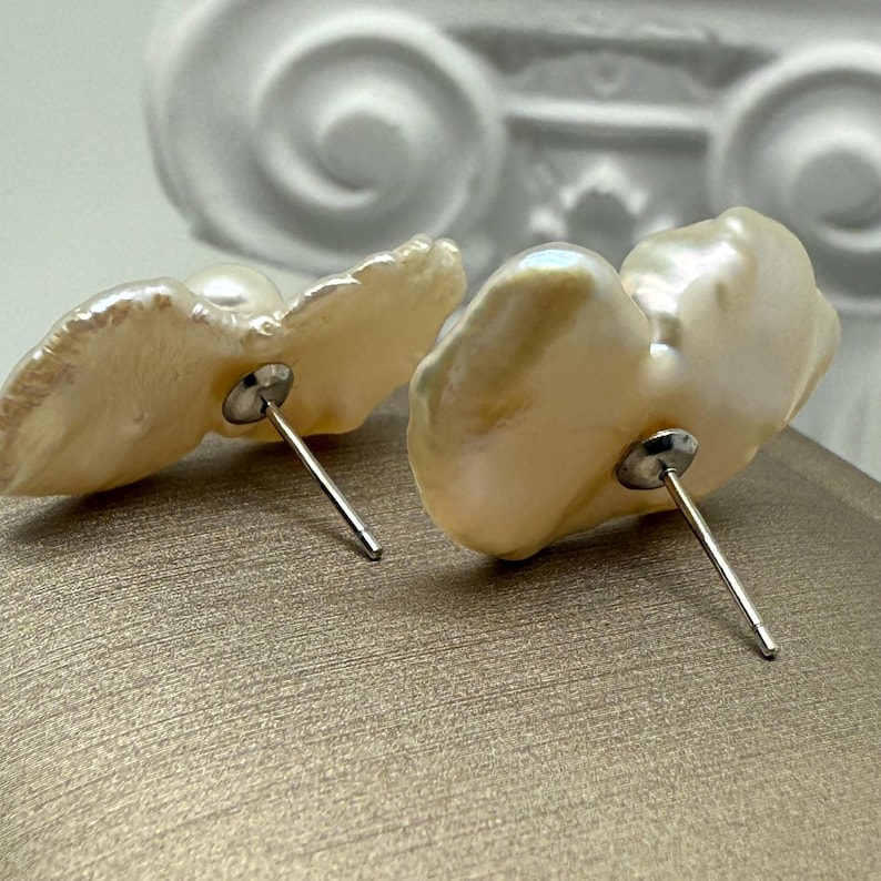 May include: A pair of pearl stud earrings. The earrings feature irregular, off-white pearls with a lustrous sheen. Each pearl is mounted on a silver post. The background is a neutral, textured surface.