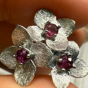 May include: A silver flower pendant featuring three pink gemstones. The pendant has a textured finish.