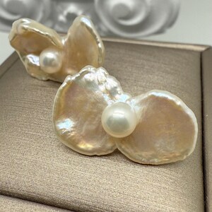 May include: Two baroque pearl earrings. Each earring features a large, iridescent, petal-shaped pearl with a smaller, round pearl at its center. The pearls have a creamy, off-white color with hints of gold and pink.