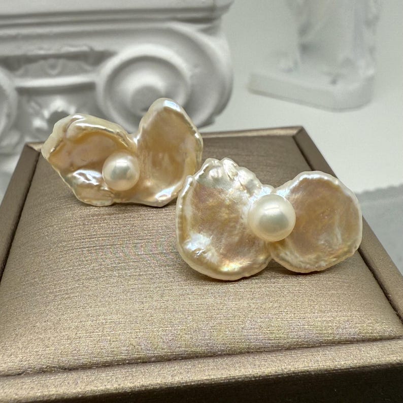 May include: A pair of pearl earrings. Each earring features a large, irregular, cream-colored pearl with a smaller, round, white pearl in the center. The earrings are displayed on a gold-toned surface.