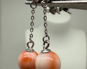 May include: A pair of drop earrings featuring round, smooth, peach-colored beads suspended from silver-toned metal chains. Each earring has a small silver-toned stud top and a delicate link chain connecting the stud to the spherical bead at the base.