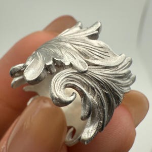 May include: A silver ring with an elaborate, leaf-inspired design. The ring showcases intricate, swirling patterns and textures, giving a three-dimensional appearance. The ring is held by a person.