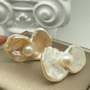 May include: A pair of pearl earrings. Each earring features a lustrous, off-white pearl set within a delicate, shell-shaped design. The shells have a creamy, iridescent sheen, creating a natural and elegant aesthetic.