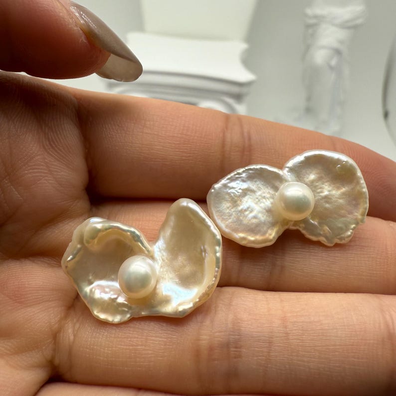 May include: Two pearl earrings. Each earring features a lustrous, off-white, petal-shaped design with a small, round pearl at the center. The earrings are set against a soft, neutral background, highlighting their delicate beauty.