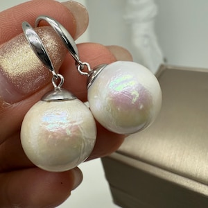 May include: A pair of white pearl earrings with silver hooks. The pearls have an iridescent sheen, reflecting pink and green hues. The earrings are round and have a simple, elegant design.