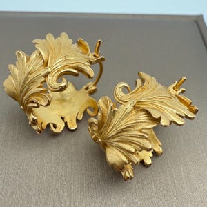 May include: A pair of gold-coloured earrings with a leaf design. The earrings feature intricate detailing, with each leaf having textured veins and a curved shape. The earrings are displayed on a neutral background, highlighting their metallic sheen.